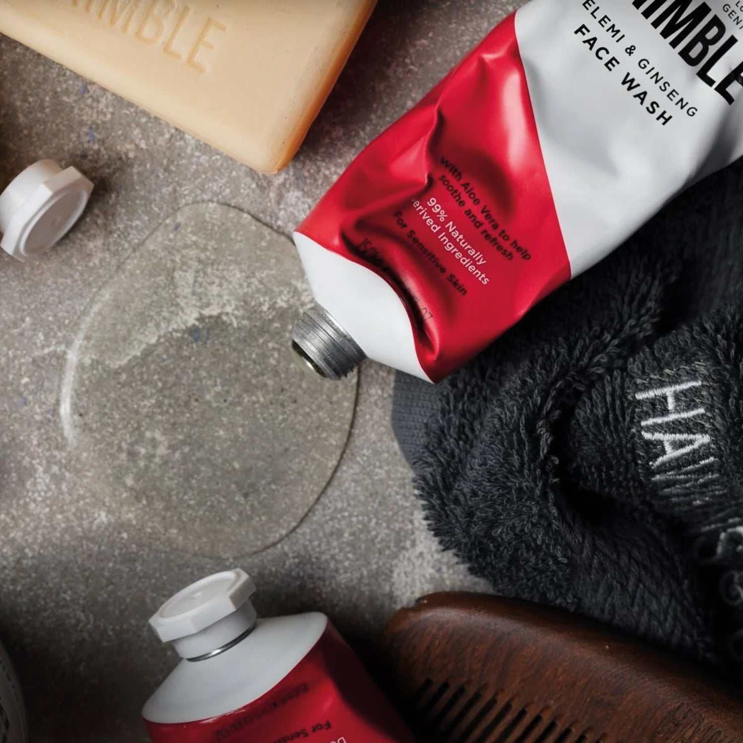 Gesichtswaschgel von Hawkins & Brimble in roter Aluminiumtube mit klarem Waschgel, Handtuch und Kamm auf grauem Untergrund