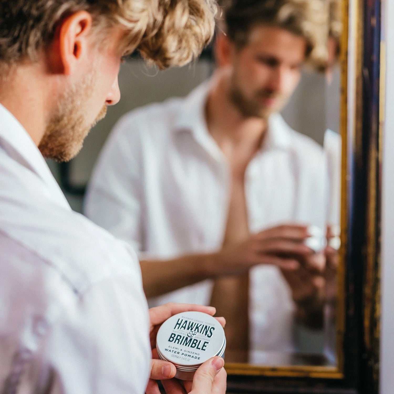 Mann hält wasserbasierte Haarpomade von Hawkins & Brimble vor Spiegel für stylisches Haarstyling