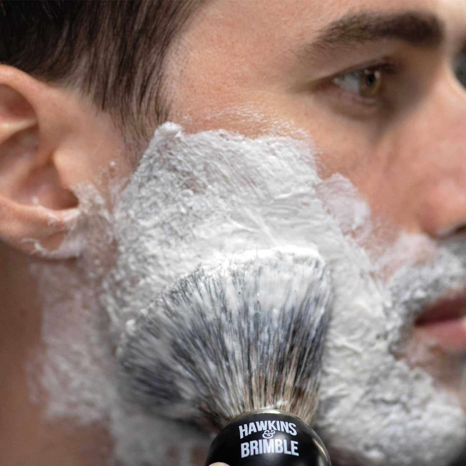 Mann trägt Rasiercreme Hawkins & Brimble mit Pinsel für sanfte und gründliche Rasur auf Gesicht auf.
